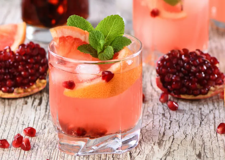Ein Glas mit einem alkoholfreien Cocktail aus Grapefruit und Granatapfel auf einem Tisch mit Spalten von Blutorangen und Granatapfel-Stücken