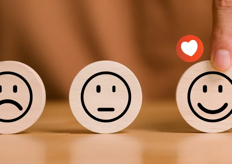 Drei Smileys zur Bewertung von Leistungen auf runden Holztäfelchen, eine Hand wählt den glücklichen Smiley aus