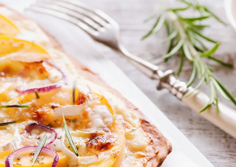 Foodpairing Rezept: Pfirsich-Pflammkuchen auf einem Brett mit Ziegenkäse, daneben eine Gabel und ein Rosmarin-Zweig