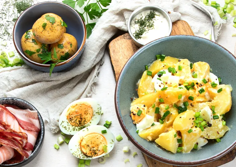 Verschiedene Sorten Kartoffelsalat in kleinen Schalen dazwischen Schälchen mit Bacon und Ei auf einer hellen Arbeitsplatte