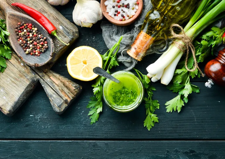 Zutaten für Chimichurri und ein Glas mit fertiger Chimichurri auf einem Holztisch