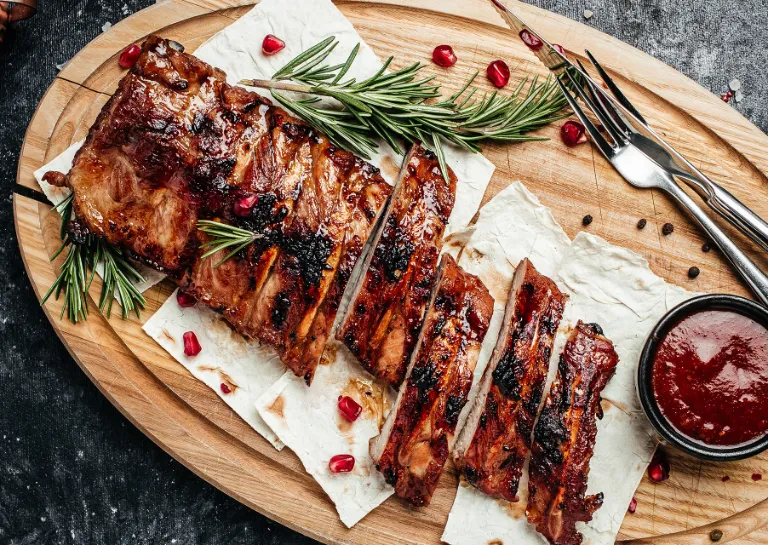 Fruchtige Schweinerippchen auf einem Holzbrett, mit Granatapfelkernen garniert, Besteck liegt dabei