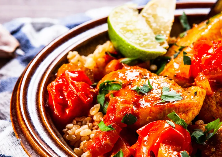 Muckalica, ein Gericht aus Schweinefleisch, Paprika und Aubergine, in einem Teller mit Limettenspalten angerichtet
