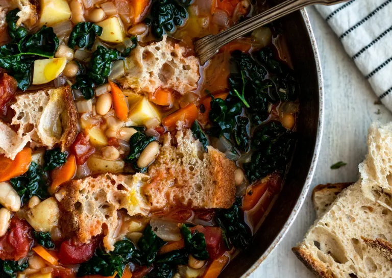 Ribollita in einem grauen Topf mit silbernem Löffel auf einem hellen Tisch, daneben liegen zwei Scheiben Brot