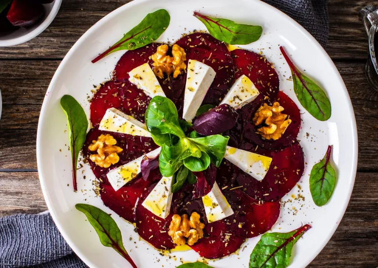 Rote-Bete-Carpaccio mit Ziegenkäse und Walnüssen auf einem weißen Teller angerichtet, daneben eine kleine Schale mit Walnüssen