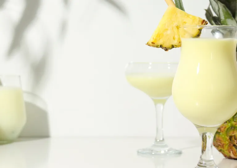 Alkoholfreier Smoothie-Cocktail mit Ananas und Kokosmilch in Cocktailgläsern auf einem weißen Tisch mit einer ganzen Ananas und zwei Kokosnuss-Hälften