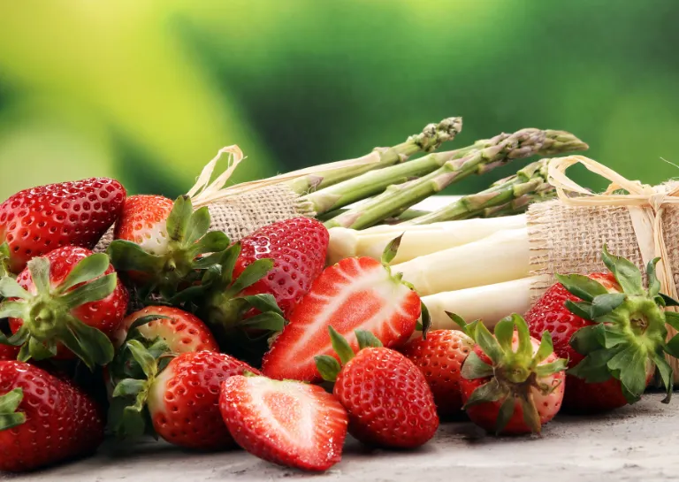 Je ein Bund grüner und weißer Spargel und frische Erdbeeren auf einem Holztresen mit grünem Hintergrund.