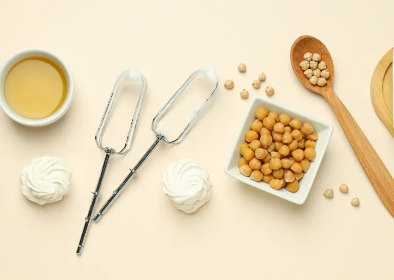 Kichererbsen in Schüsseln und auf Löffeln, Aquafaba in Schüsseln & veganer Einschnee aus Aquafaba in Schüsseln mit Schneebesen auf einem Tisch
