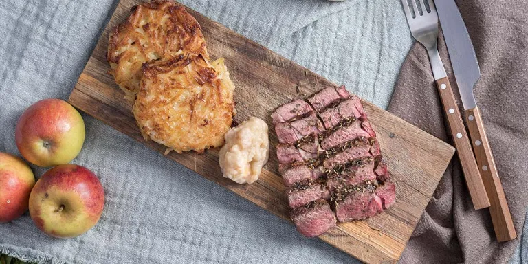 Gegrillte Rehkeule liegt mit Apfel-Zwiebel Dip und Petersilienwurzel-Kartoffel Reibekuchen auf einem Holzbrett