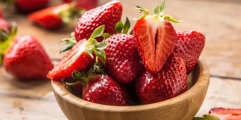 Frische Erdbeeren in einer Holzschale und dahinter auf einem Holztisch verteilt.