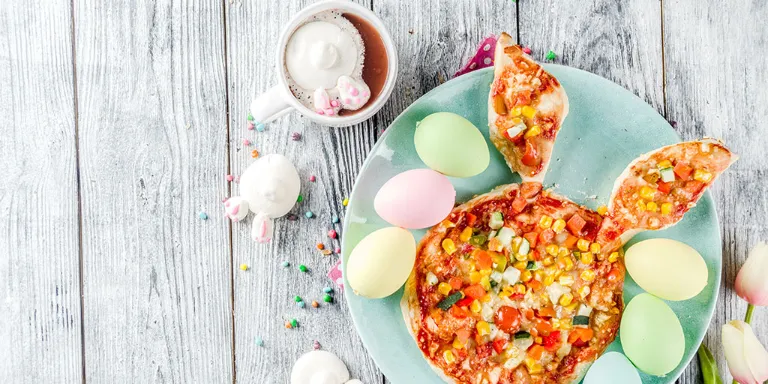 Frisch gebackene Pizza in Hasenform angerichtet mit bunten Ostereiern auf einem pastellblauen Teller auf einem Holzuntergrund. 