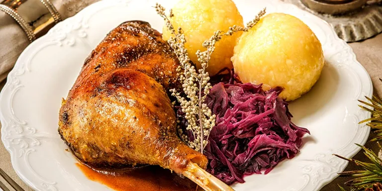 Keule von der Martinsgans angerichtet auf einem weißen Teller mit Knödeln und Rotkohl. Im Hintergrund Kerzenschein und eine Sauce sowie Weihnachtsdeko