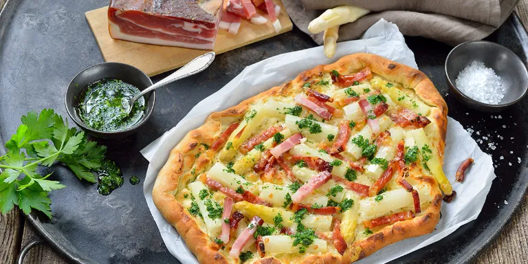 Frisch gebackener Flammkuchen mit weißem Spargel und Schinken auf einer schwarzen Servierplatte angerichtet mit frischem Pesto.