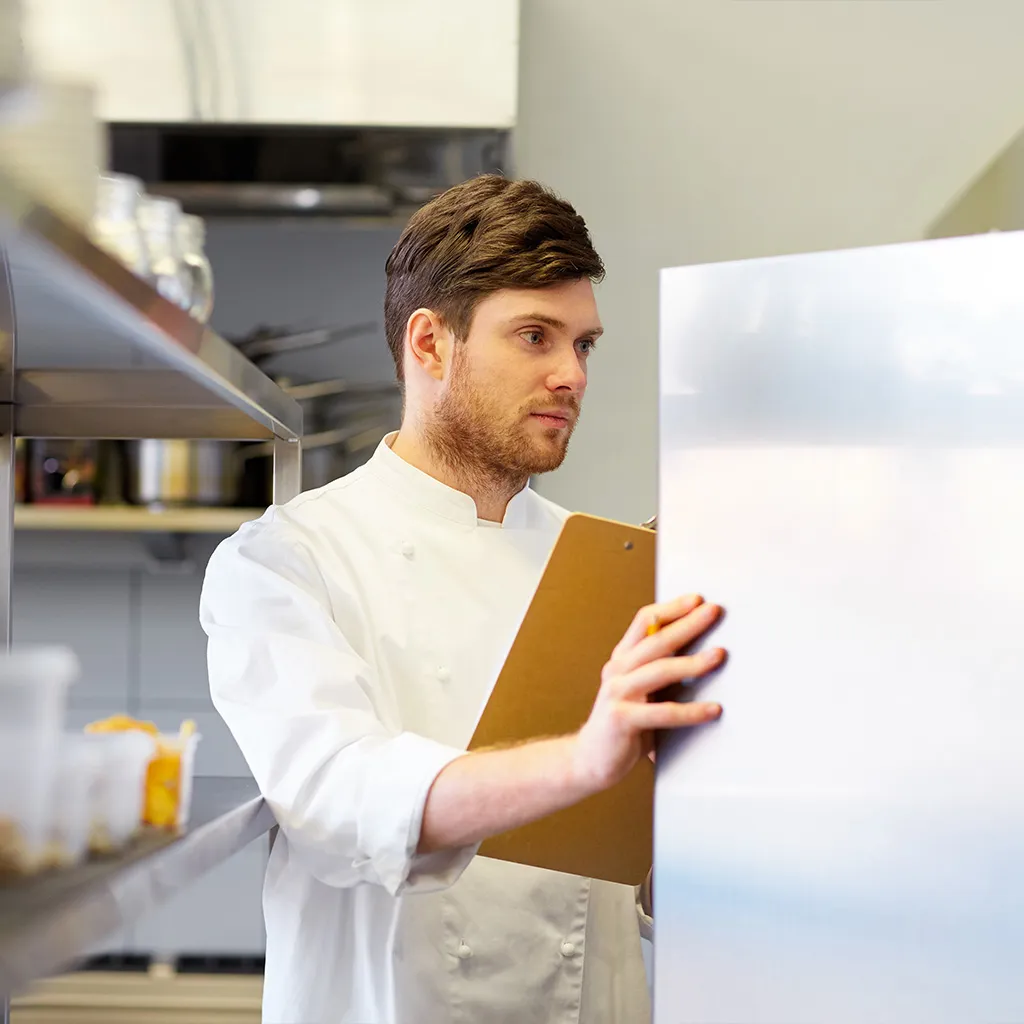 Koch schaut in Kühlschrank und hält eine Checkliste in der Hand