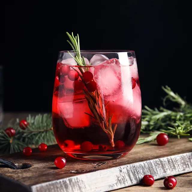 Ein Glas mit einem alkoholfreien Cocktail aus Cranberry und mit Rosmarin auf einem Holzbrett vor dunklem Hintergrund, dekoriert mit Tannenzweigen, Rosmarin und Cranberrys