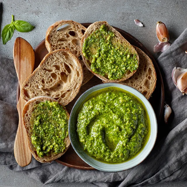 Grünes Pesto in einer grauen Schale mit Brotscheiben auf einem Brett angerichtet