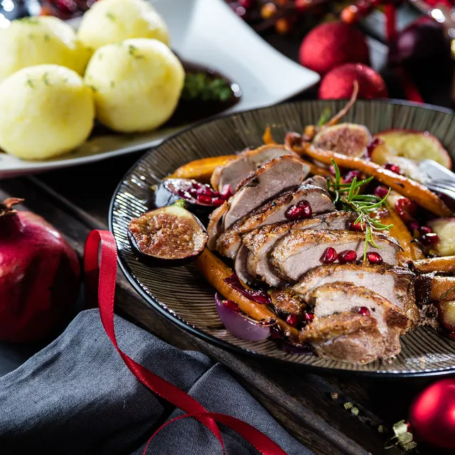Entenbrust, aufgeschnitten und auf einen dunkelgrauen Teller mit Granatapfelkernen angerichtet, mit Klößen serviert