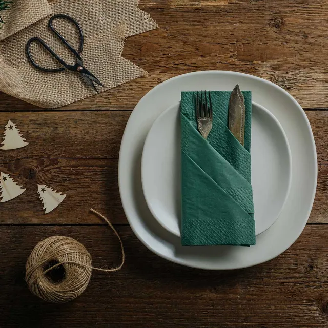 Grüner Bestecktasche aus einer Serviette gefaltet auf einem weißen Teller mit Besteck. Auf einem Holztisch mit Garn und Schere. 