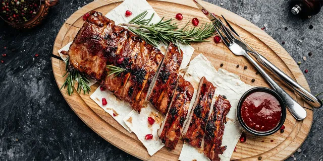 Fruchtige Schweinerippchen auf einem Holzbrett, mit Granatapfelkernen garniert, Besteck liegt dabei