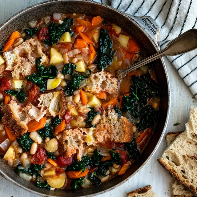 Ribollita in einem grauen Topf mit silbernem Löffel auf einem hellen Tisch, daneben liegen zwei Scheiben Brot