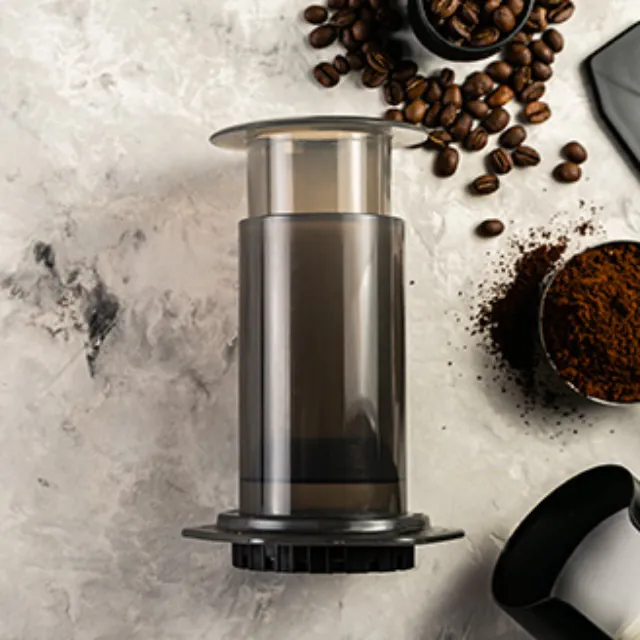 Eine AeroPress zur Zubereitung von Kaffee liegt auf einem grau marmorierten Tisch. Daneben sieht man Kaffeebohnen und Kaffeepulver liegen