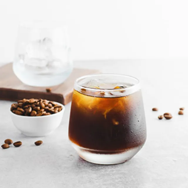 Cold Brew-Kaffee in einem Glas mit Eiswürfeln, dahinter sieht man eine kleine Schale mit Kaffeebohnen