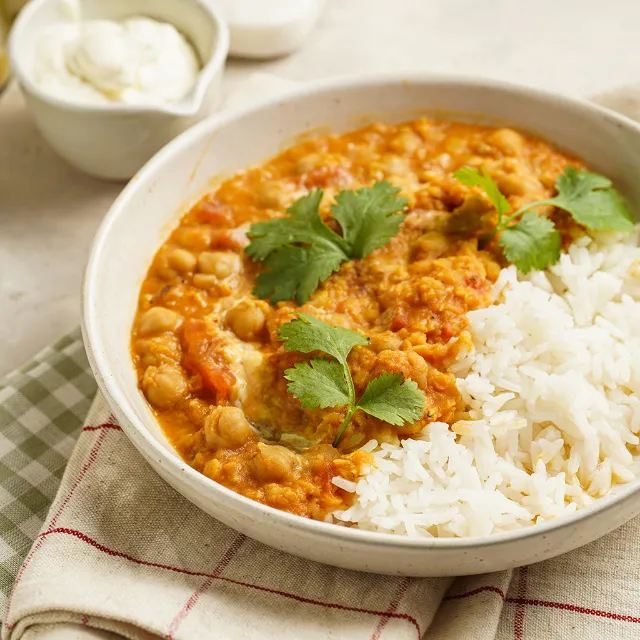 Curry mit Kichererbsen angerichtet mit Reis in einer weißen Schüssel auf einem weiß-rot-karierten und einem grün-weiß-karierten Küchentuch auf einem Tisch