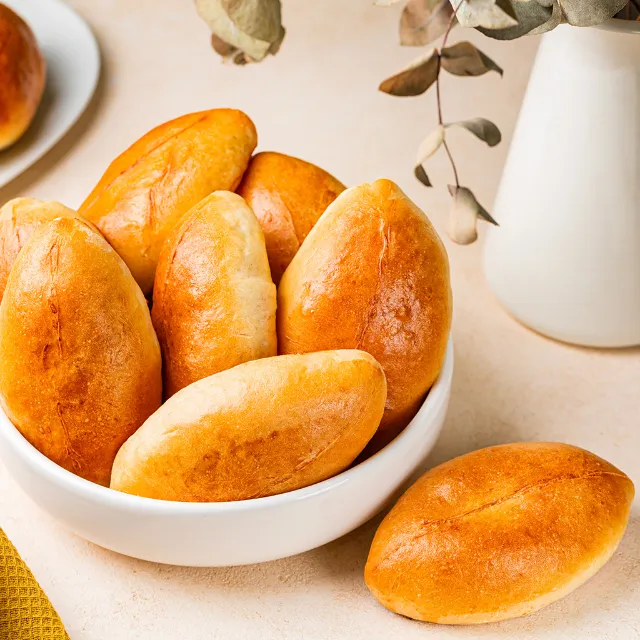 Sechs ovale Brötchen aus süßem Hefeteig in einer weißen Schüssel auf einem Tisch, daneben liegt ein weiteres Brötchen