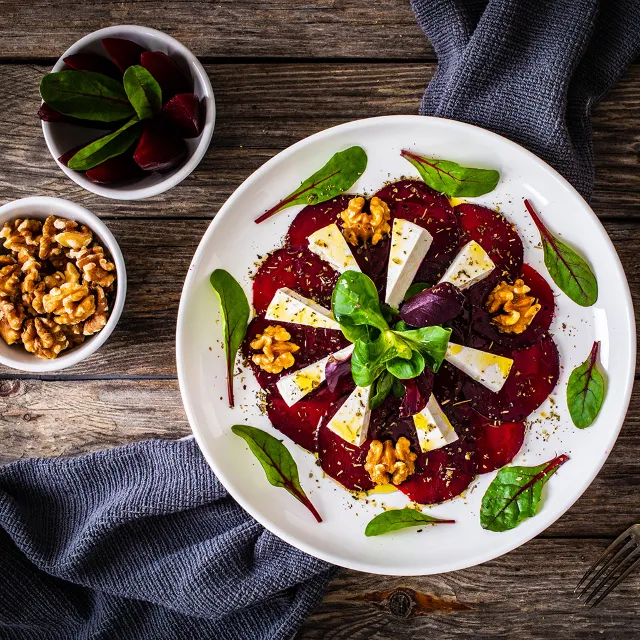Rote Bete Carpaccio mit Ziegenkäse und Walnüssen auf einem weißen Teller angerichtet, daneben Schüsselchen mit Walnüssen und Rote Bete Würfel