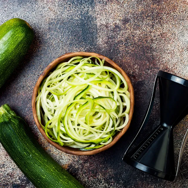 Zucchini-Zoodles liegen in einer Schüssel, daneben ein Zucchini-Spiralschäler und ganze grüne Zucchinis